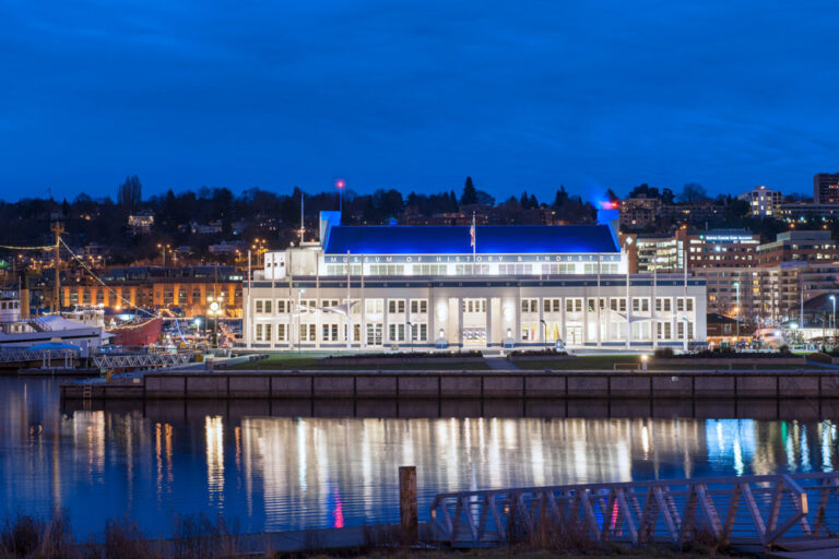 Against a backdrop of city lights at dusk, a waterfront building with bright white lights offers a captivating view—a must-see among things to do in Seattle. A boat is moored nearby, while the deep blue sky and water reflect the luminous scene perfectly.