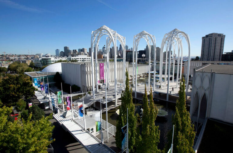 Aerial view of a futuristic building with tall, white arch structures in the vibrant cityscape of Seattle. Surrounded by lush trees and a stunning skyline under a clear blue sky, it's one of the best things to do in Seattle for architecture lovers.