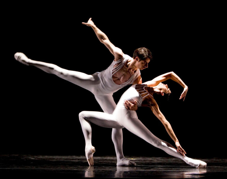 In a mesmerizing display, two ballet dancers in white costumes captivate the audience on stage, silhouetted against a black backdrop. As part of the must-see things to do in Seattle, the male dancer lifts his partner effortlessly, her arm extended gracefully in an elegant pose.