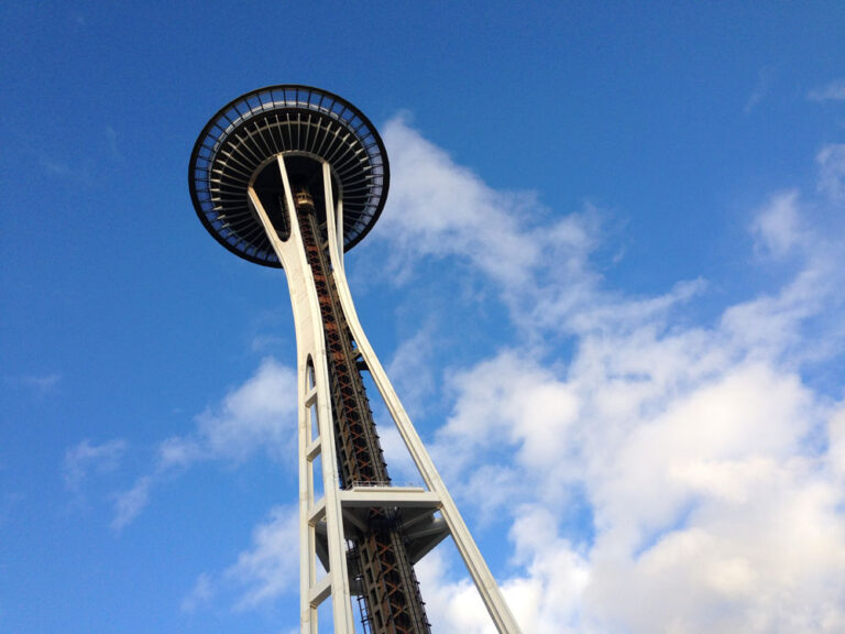 View of the Space Needle from below against a bright blue sky with white clouds. Among the top things to do in Seattle, this iconic structure towers above, showcasing its unique design and observation deck.