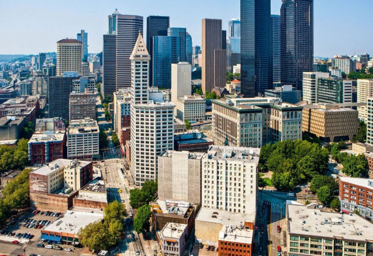 Aerial view of a bustling cityscape featuring a mix of modern skyscrapers and historic buildings. Streets lined with trees weave through the urban environment, echoing the vibrant essence of things to do in Seattle, while distant hills are visible under a clear blue sky.