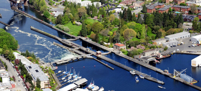 Aerial view of a canal with boat locks, reminiscent of those found among the things to do in Seattle, surrounded by greenery and industrial buildings. Boats are docked nearby, trees are scattered across the area, and a residential neighborhood forms the picturesque backdrop.