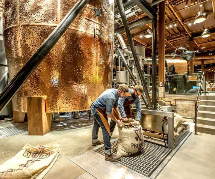 Two workers in aprons handle large burlap sacks inside a distillery near large copper tanks and industrial equipment. This modern space, with exposed beams and warm lighting, is a hidden gem among the things to do in Seattle for those seeking unique local experiences.