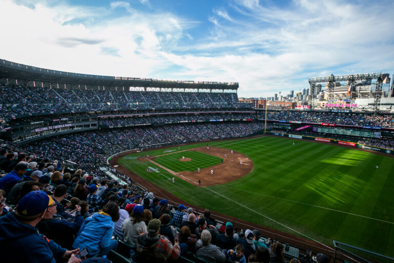 Experience one of the top things to do in Seattle with a wide-angle view of a bustling baseball stadium during a daytime game. The stands are packed with fans, and the city skyline graces the background under a partly cloudy sky, creating an unforgettable scene.