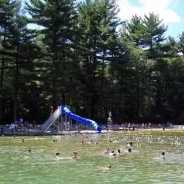 Filtered swimming facility with sandy beach, water slide, and people swimming, surrounded by trees.