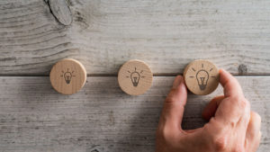 hand placing three wooden cut circles with light bulb icon drawn on them over textured white wooden background.