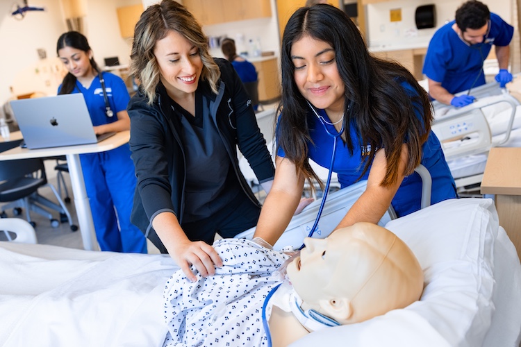 clinical assistant professor helps a nursing student use a stethescope on a manikin in the simulation center