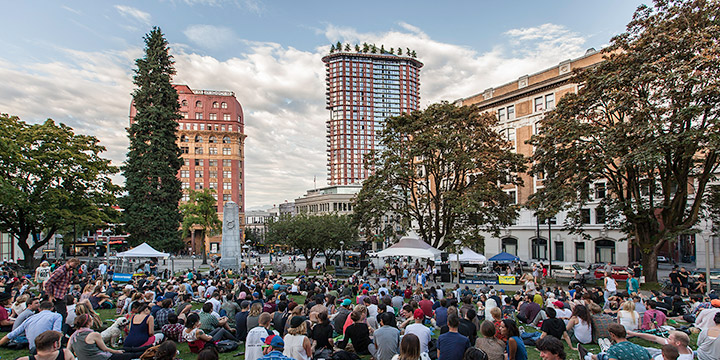 People gathered at Victory Square to watch a music concert, with heritage buildings along Hastings Street in the background