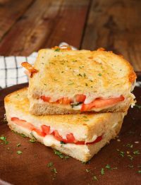 a vertically aligned photo of two halves of a Caprese Grilled Cheese that have been stacked one atop the other on a dark wooden serving tray with a black and white stripped napkin visible in the background