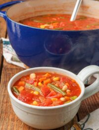 tomato based soup with corn, green beans, and great northern beans in a blue enamel soup pot with a white soup bowl full