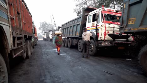 Woman-carrying-coal,-Mine-trucks-used-for-coal-shipment-waiting-for-loading-inside-Jharia-coalfield,-Dhanbad,-Jharkhand