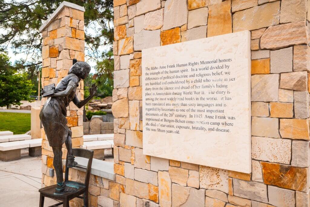 A statue of Anne Frank balancing on a chair looks out a window next to an interpretive sign.