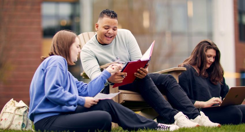 Three people sit on the grass, one holding a red binder, deep in discussion. They appear to be students from Worcester State University, perhaps collaborating on a project or study session.