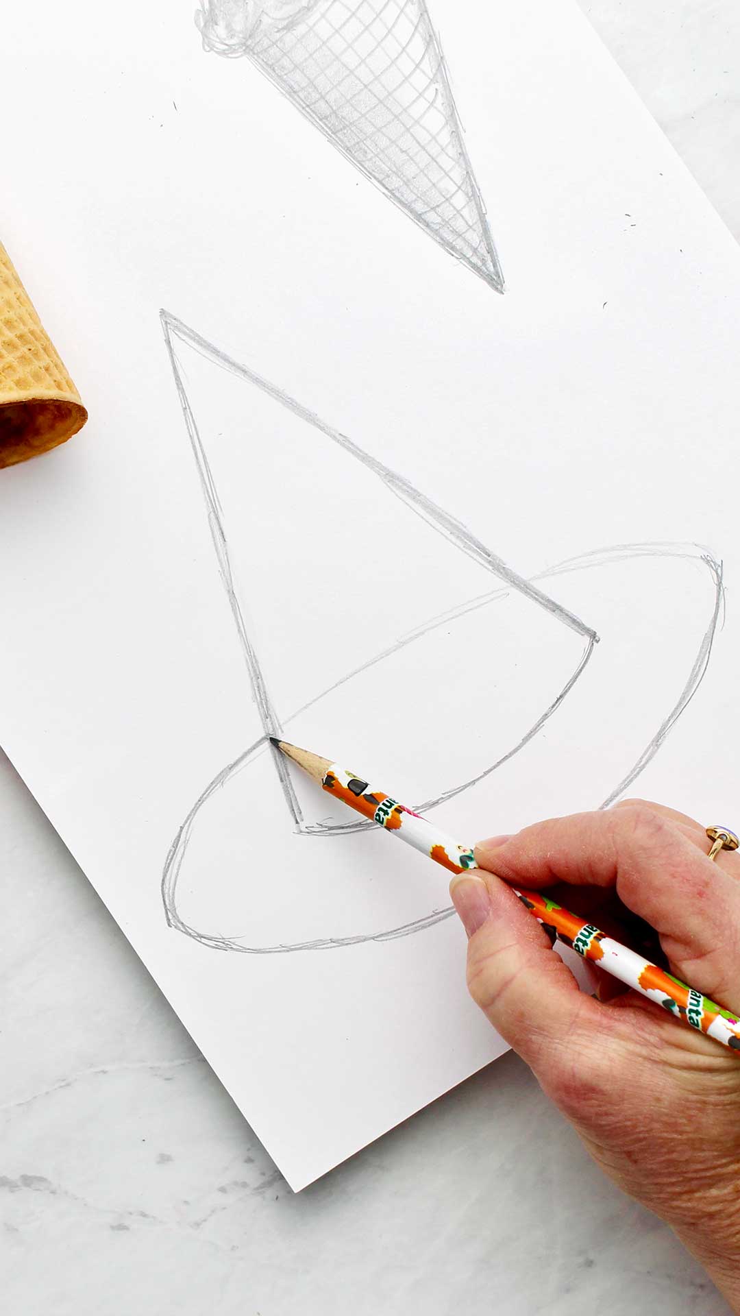 A hand sketching an oval for the brim of the witches hat drawing.