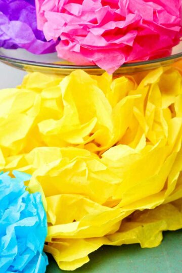 Completed colorful tissue paper flowers, a purple and pink one resting on a glass cake stand with others underneath.