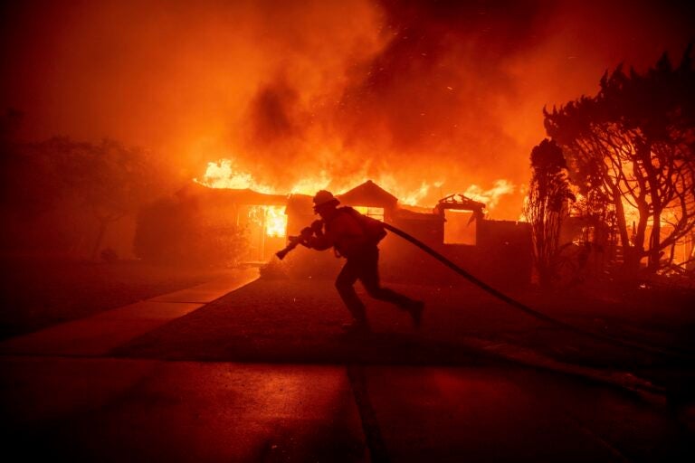 A firefighter dragging a fire hose in front of a house completely engulfed in flames