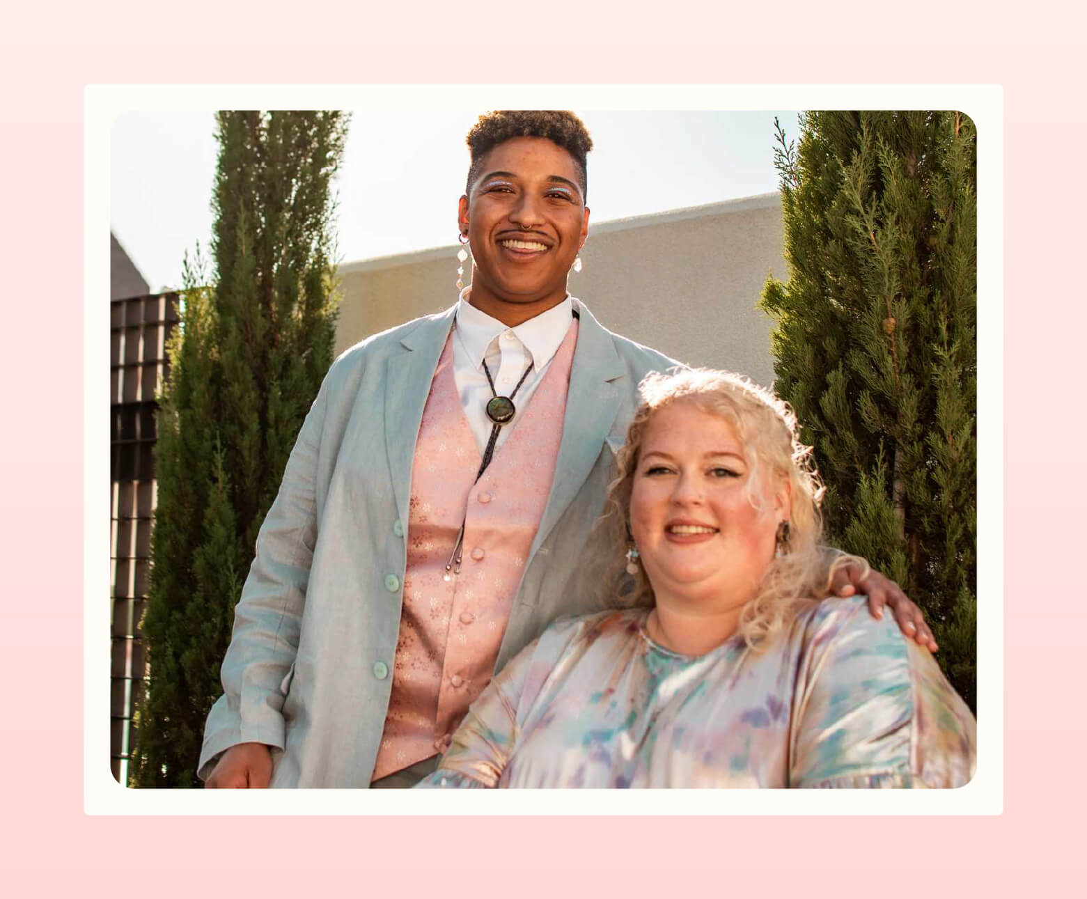 A couple poses for camera, the one standing wearing a Kirrin Finch suit with vest and bolo tie and the one seated wearing a colorful dress