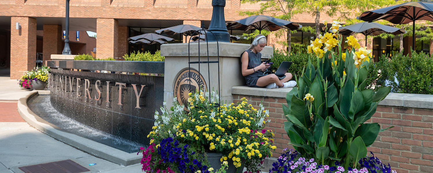 A Kent State University student types on her phone while sitting with a computer by the fountain on Risman Plaza. (Photo credit: Bob Christy)