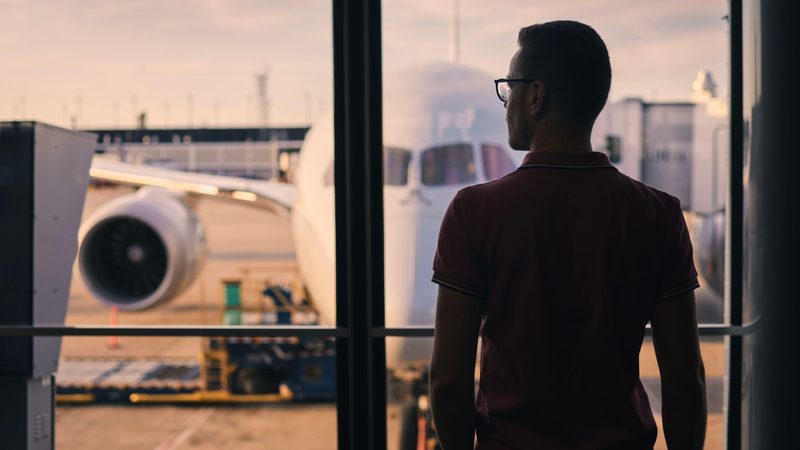 Traveller is looking out of airport window at airplane. Silhouette of man waiting for his flight