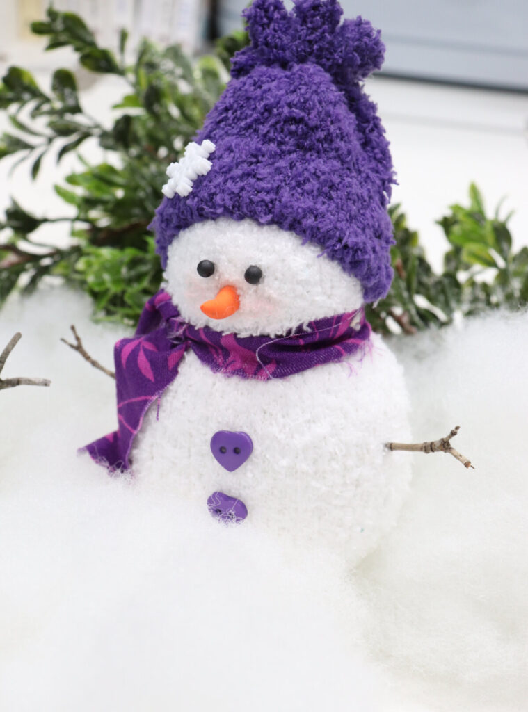 Image contains a sock snowman with a purple hat and scarf, sitting on a white table with Poly-Fil and greenery.