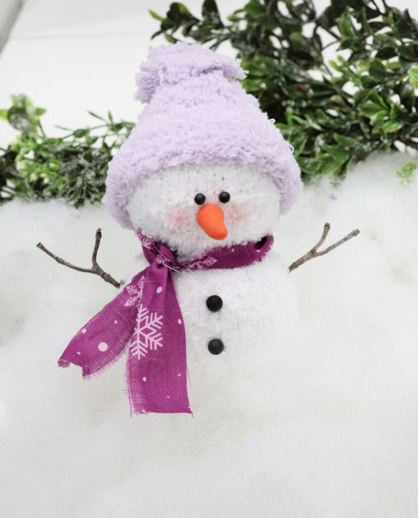 Image contains a sock snowman with a purple hat and scarf and black buttons. It sits on a pile of Poly-Fil on a white table, with greenery in the background.