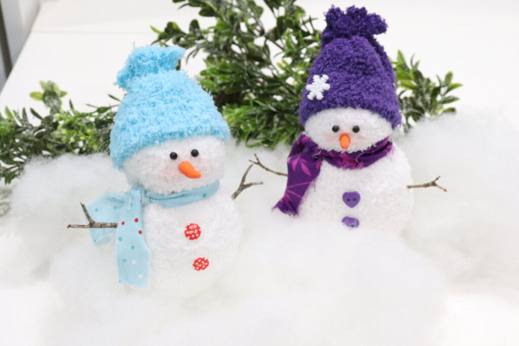 Image contains two sock snowmen; one with a blue hat and scarf, and one with a purple hat and scarf. They sit on a white table with Poly-Fil and greenery.