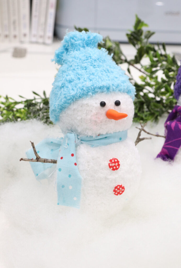 Image contains a plush snowman made from a white fuzzy sock, with a blue hat and scarf, and red buttons. It sits on a white table with faux greenery.