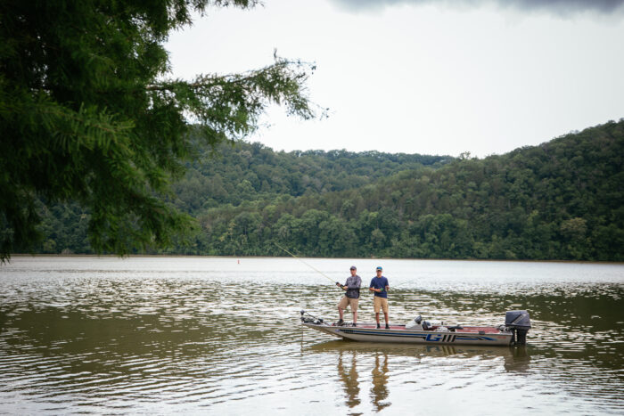 Two men stand in boat with fishing poles surrounded by mountains