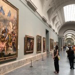 Masked museum visitors gaze at paintings hung in a long hallway.