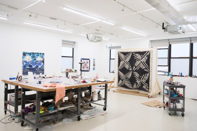 View of an artist studio with a word bench at left and a quilt hanging on a wooden structure at center. 