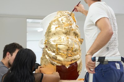 Three studio assistants apply gold lef to a marble bust. 