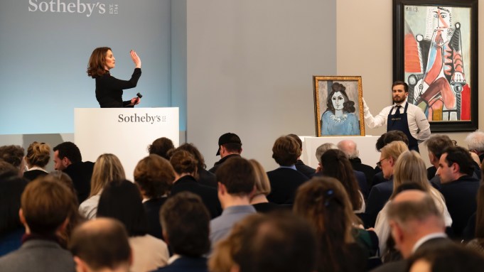 A woman standing behind a dais gestures at a crowd beside two paintings.