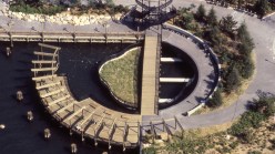 An overview view of a circular land arrangement jutting out into water, with rounded walkways and a black tower to climb.