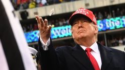 A man in a suit and a red hat tosses a coin at a stadium. 