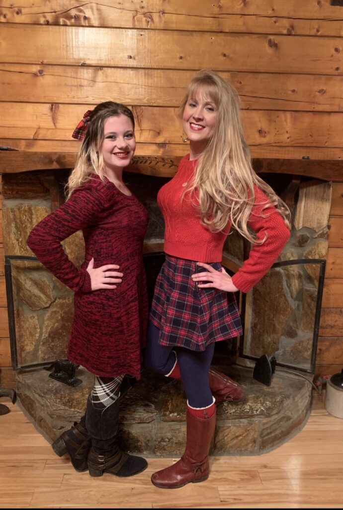 Photo of two blonde, White women standing side-by-side and wearing red outfits. They are smiling at the camera.