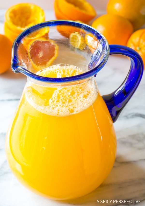 Orangeade in a glass pitcher with oranges in the background. 