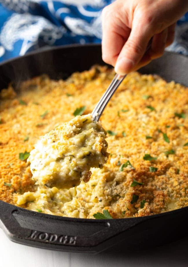 Hand with metal spoon scooping out portion of casserole from a cast iron skillet.