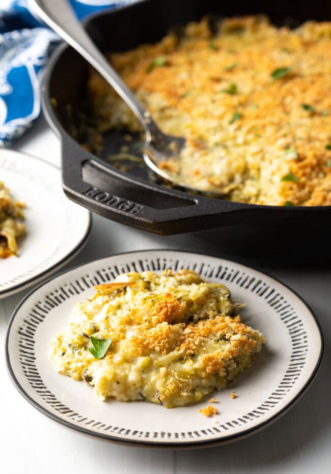 Plate with zucchini casserole serving on it. Cast iron with casserole in background.