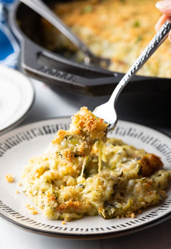 Fork holding bite of cheesy zucchini casserole from the plate to camera.