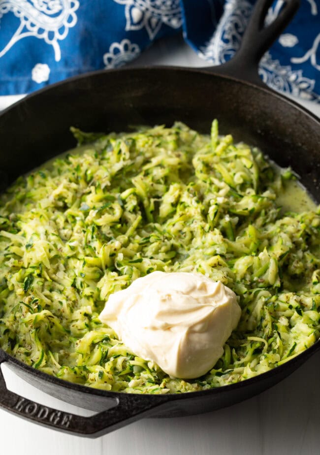 Grated zucchini in a cast iron skillet, large dollop of mayonnaise on top of the zucchini.