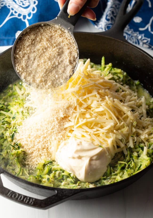 Hand with measuring cup adding Panko breadcrumbs to the skillet with zucchini, mayo, and shredded cheese.