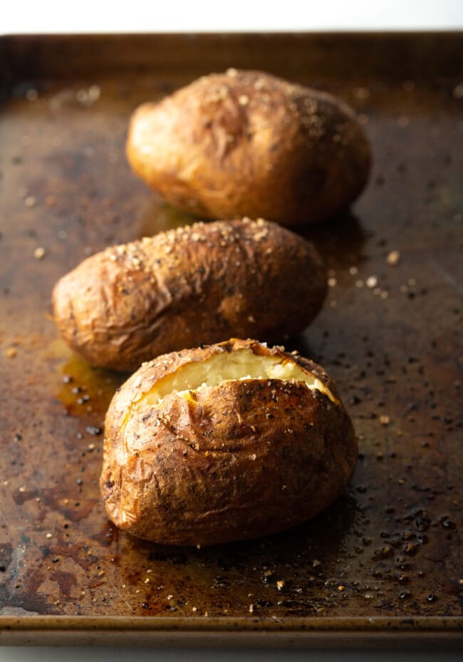 Three baked potatoes on a baking sheet. One closest to camera is split open at the top.