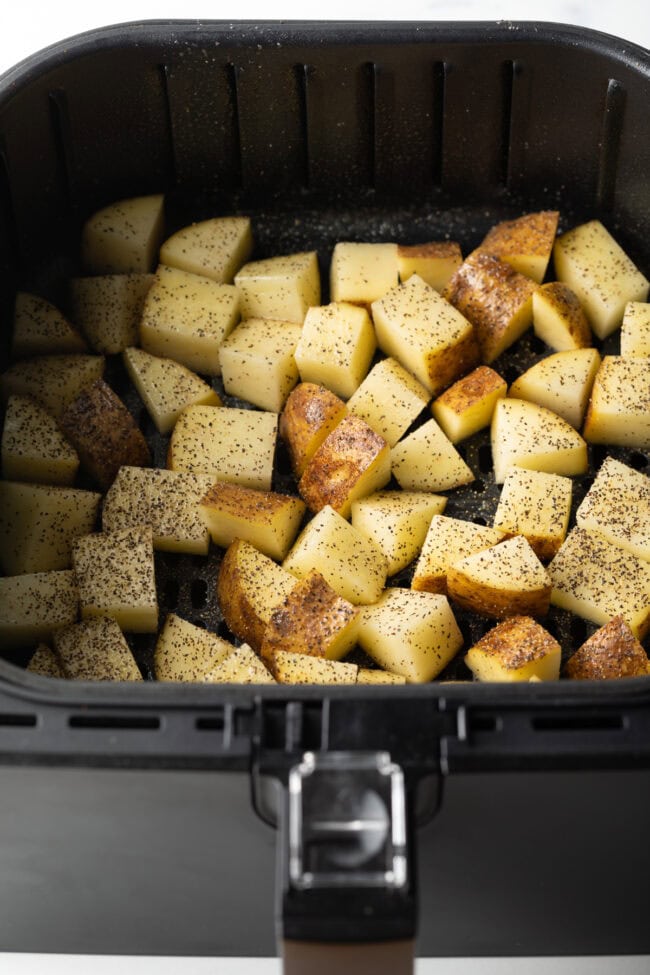 Raw pieces of cubed potato in the basket of an air fryer, seasoned with salt and pepper.