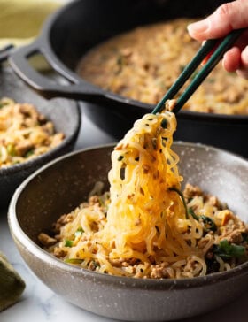 Spicy Szechuan noodles in a black bowl topped with green onion slices. Hand with set of chopsticks are grabbing a bite of noodle.