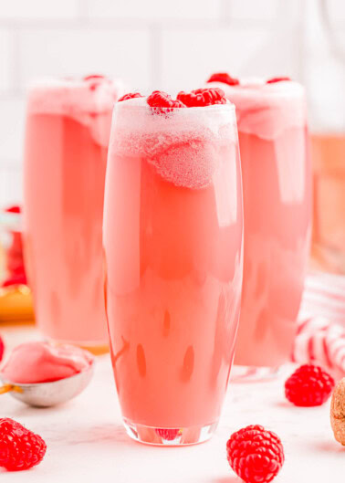 Three tall glasses filled with a frothy pink beverage, topped with raspberries, surrounded by scattered raspberries and a glimpse of a spoon with a pink substance.