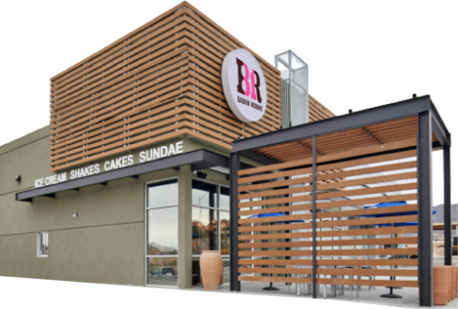 A modern Baskin Robbins storefront with a wooden slat facade, large windows, and an outdoor seating area under a pergola. Signs for dessert and ice cream franchise offerings like shakes, cakes, and sundaes are visible.