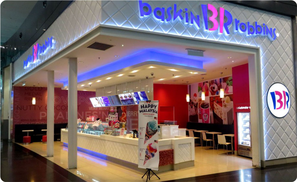 A Baskin Robbins store with a white and pink interior, displaying ice cream tubs. There's a "Happy Malaysia" promotional banner near the entrance, celebrating their renowned dessert and ice cream franchise.