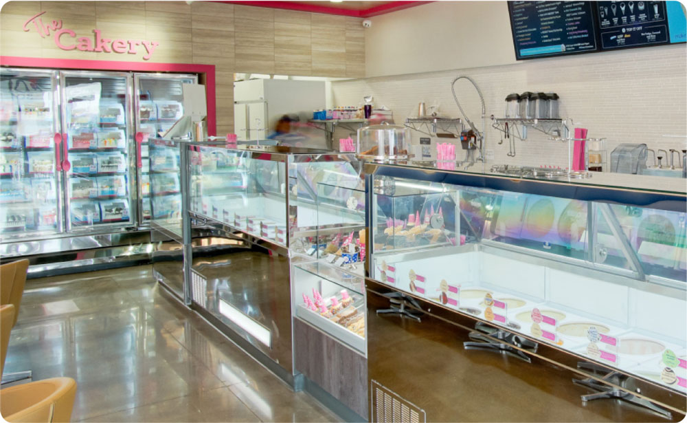 A brightly lit bakery with pink decor, display cases filled with various pastries, cakes, and gelato from a popular dessert franchise, and a large menu board overhead. A person is visible behind the counter.