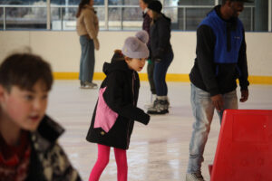 A young girl enjoys ice skating at the Pavilion with other patrons around her.