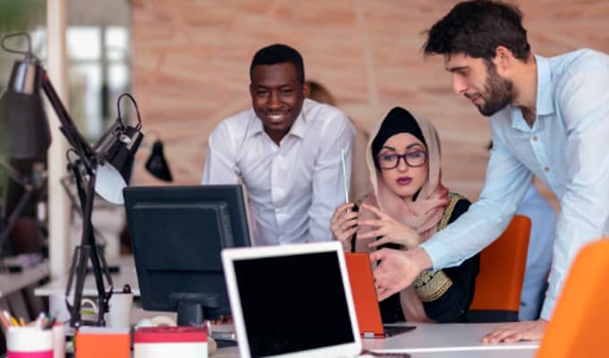 Three colleagues collaborate at a shared desk in an office setting. Two men and a woman in a hijab are engaged in a discussion over a laptop. Office equipment and decor are visible in the background.